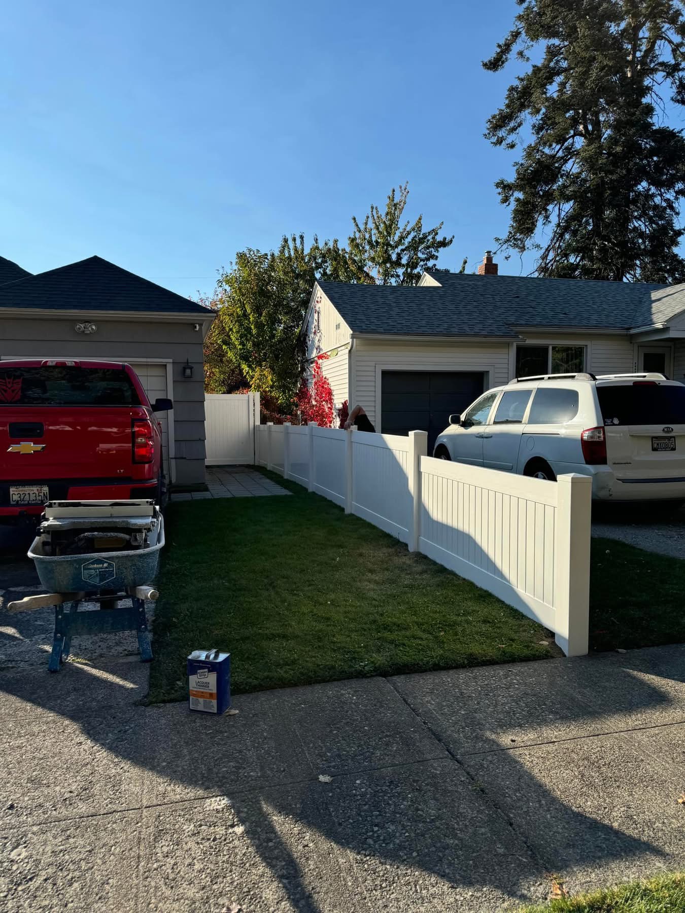 Robertson Fence co in Spokane, WA A white vinyl fence runs between two suburban houses, separating their driveways. A red truck and a white SUV are parked, with tools and paint supplies on the sidewalk and driveway. Trees and blue sky are in the background.